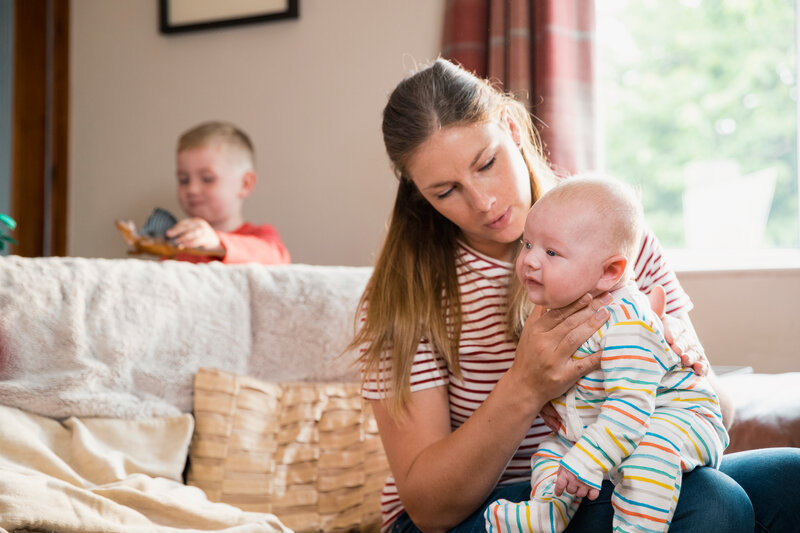 A mother is pictured holding her baby sat on a sofa while another child plays behind them